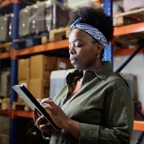 A worker in a warehouse reviews a tablet while standing near shelving stocked with boxes