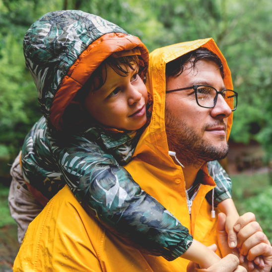 Person in a yellow rain jacket carrying a child on their back outdoors among green trees.