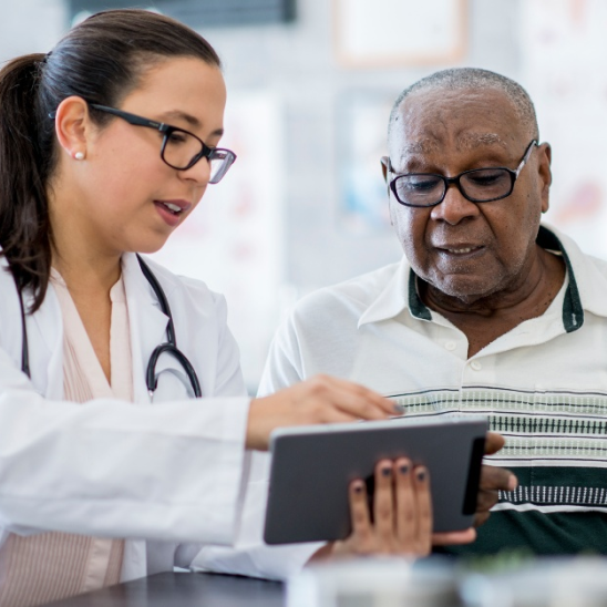 A healthcare provider in a white coat and stethoscope reviews a tablet with a person wearing glasses