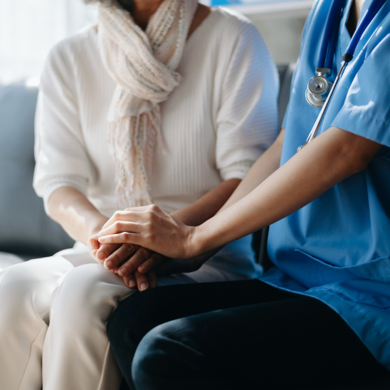 A healthcare provider in blue scrubs and stethoscope gently holds the hands of a seated person in a white sweater