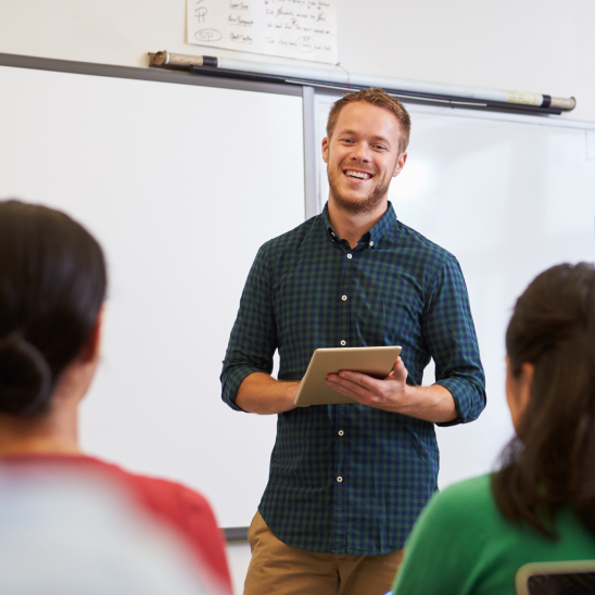 Smiling teacher holds a tablet at a whiteboard while two people face them