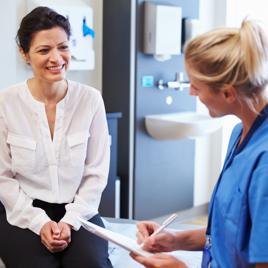 Person in a white blouse speaks with a healthcare provider in blue scrubs holding a clipboard in a clinic