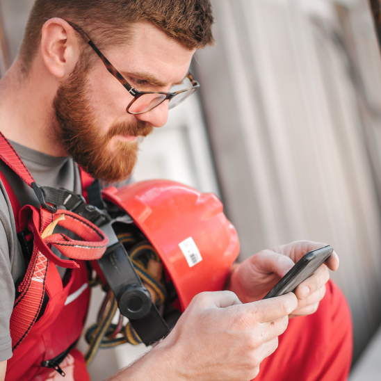 Person wearing glasses and a red hard hat looks down at a smartphone at a worksite