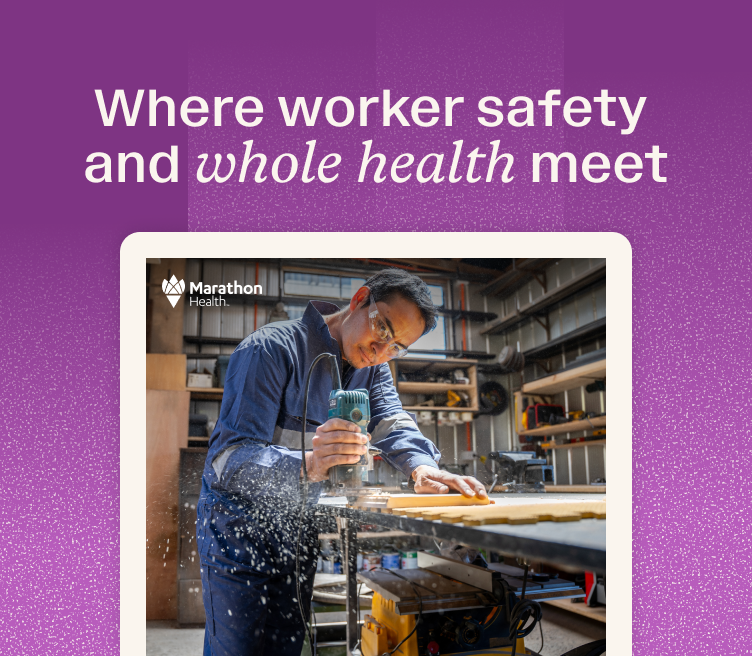 A person in blue coveralls and safety glasses uses a power tool on wood in a workshop with sawdust flying