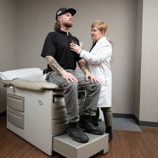 A healthcare provider in a white coat examines a tattooed worker seated on an exam table in a clinical room