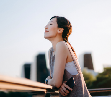 Person leans on a railing with eyes closed in warm sunlight against a city skyline