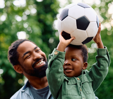 Smiling adult watches a child balance a soccer ball overhead among green trees
