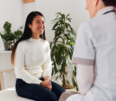 Smiling person in a white sweater sits on an exam table talking with a provider near potted plants