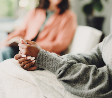 Person in a gray sweater clasps hands while talking with another person seated across the room