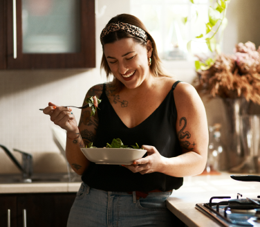 Smiling person in a leopard-print headband eats a bowl of salad in a kitchen