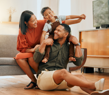 Smiling adult carries a laughing child on their shoulders while sitting with family in a living room