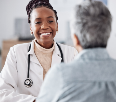 Smiling doctor in a white coat and stethoscope talks with a person across a bright office
