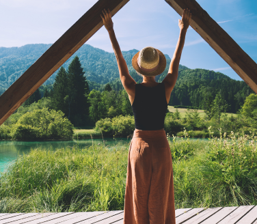 Person in a sun hat stretches arms up on a wooden deck overlooking a turquoise lake and mountains