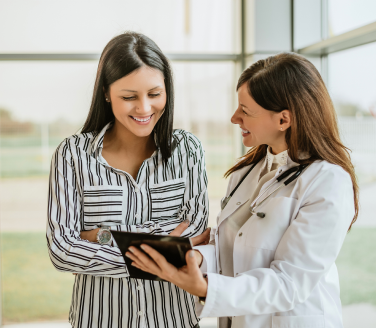 Provider in a white coat shows a tablet to a smiling person standing by tall windows