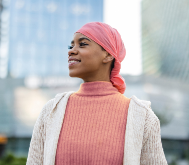 Smiling person in a pink head scarf and sweater standing outdoors among tall buildings