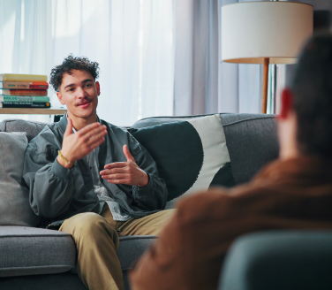 Person on a couch gestures while talking with another person in an armchair near a lamp