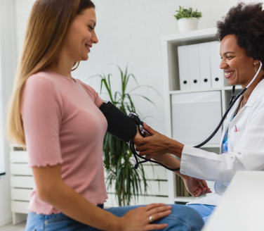 Smiling provider in a white coat takes a seated person's blood pressure in a bright office