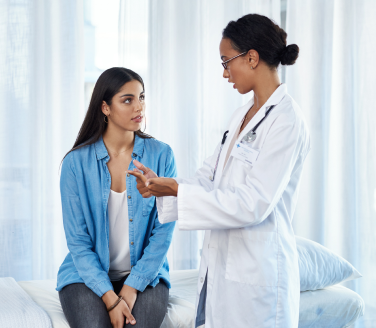 Healthcare provider in a white coat talks with a person seated on an exam bed by sheer curtains
