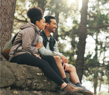 Two smiling people sit close together on a rocky ledge in a sunlit pine forest