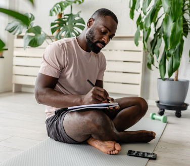 Person sitting cross-legged on an exercise mat writing in a notebook near houseplants and a dumbbell