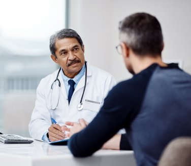 Doctor in a white coat with a stethoscope takes notes while talking with a person across a desk