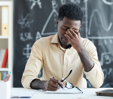 Person at a desk pinches the bridge of their nose in front of a chalkboard with diagrams