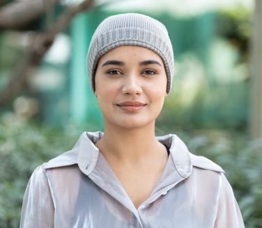 Smiling person in a gray knit beanie and shimmery blouse standing outdoors among greenery