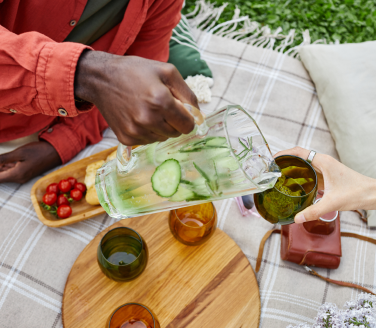Person pours cucumber water into glasses on a picnic blanket with tomatoes and flowers nearby