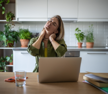Person at a kitchen table stretches their neck with eyes closed beside a laptop