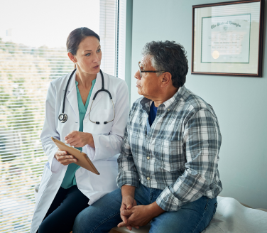 Healthcare provider with a tablet talks with a person in a plaid shirt seated in an exam room