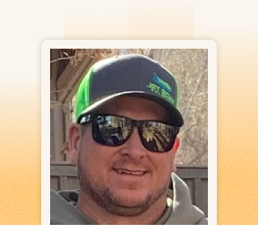 Cody smiling outdoors in a green and gray snapback cap and mirrored sunglasses near a wooden fence