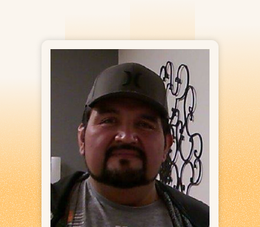 Carlos wearing a dark baseball cap and casual jacket, standing indoors near a decorative metal wall piece