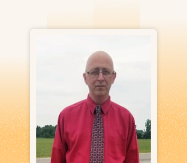 Brian Wiley standing outdoors in a red dress shirt and patterned tie, with green fields and cloudy sky behind him