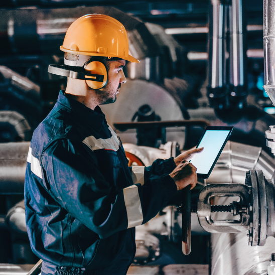 A worker in an orange hard hat and ear protection uses a tablet in an industrial plant