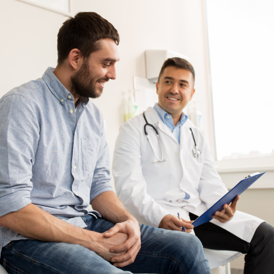 A provider in a white coat holds a clipboard while talking with a smiling person in an exam room
