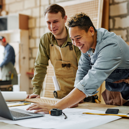 Two smiling people review plans on a laptop at a woodworking bench