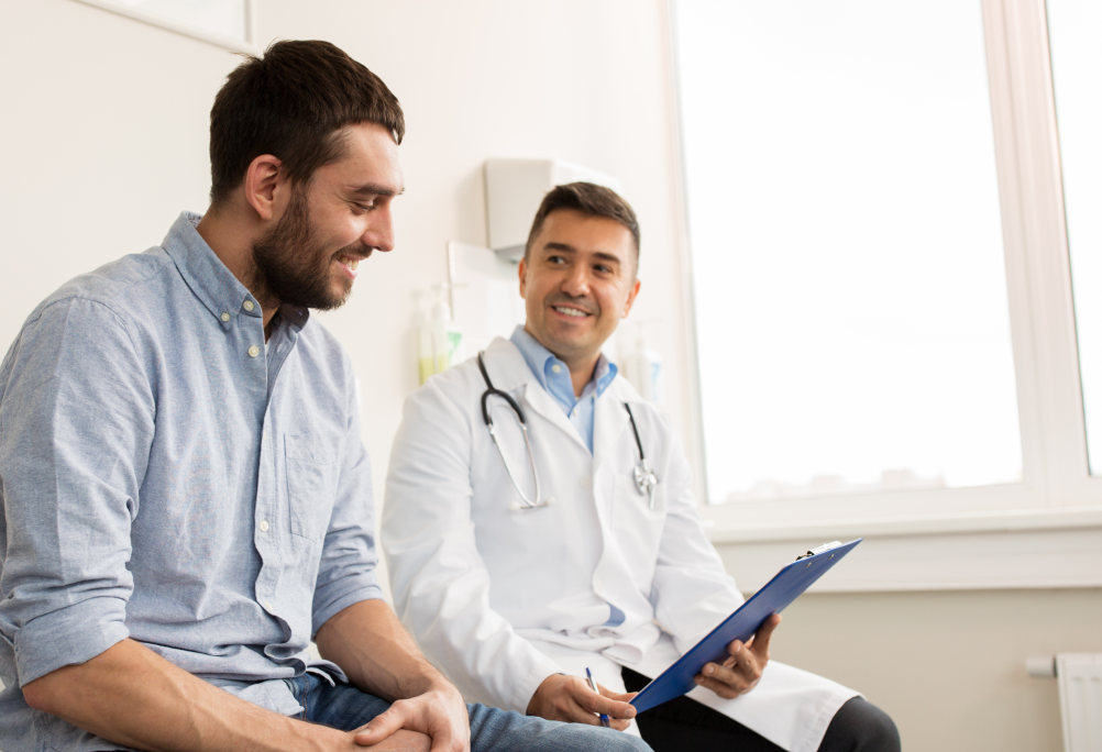 A provider in a white coat holds a clipboard while talking with a smiling person in an exam room