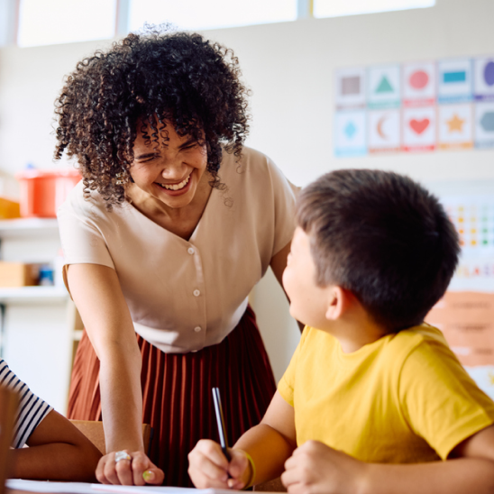 An adult laughs with a child writing at a desk in a classroom