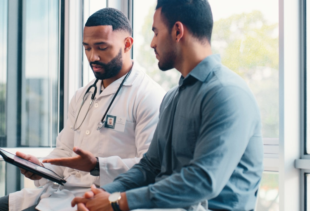 A provider in a white coat shows a tablet to a seated person in an exam room