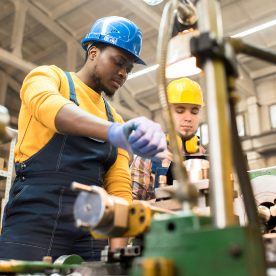 Two workers in hard hats operate industrial machinery, one guiding a part with gloved hands