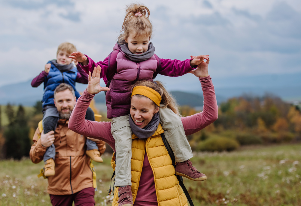 Two adults carry two children on their shoulders while walking outdoors in open countryside