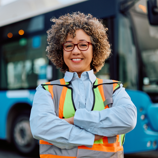 A smiling worker in glasses and a high-visibility vest standing with arms crossed in front of a bus