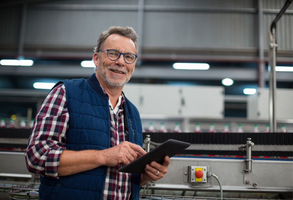 A smiling worker in glasses and a vest using a tablet beside a bottling production line